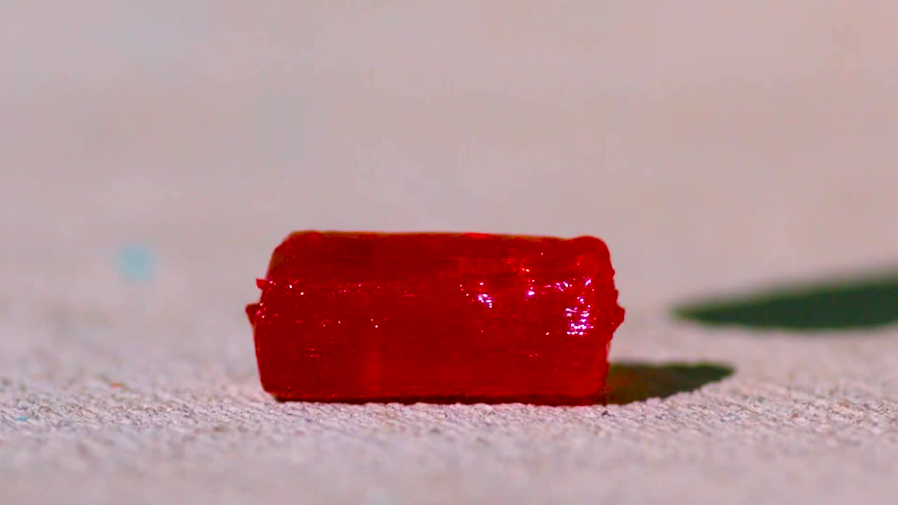 Red rock candy being crushed and broken by a hammer in slow motion. After smashing the candy it forms into hundreds of pieces moving outwards. Footage has a concrete gray background blurry background.
