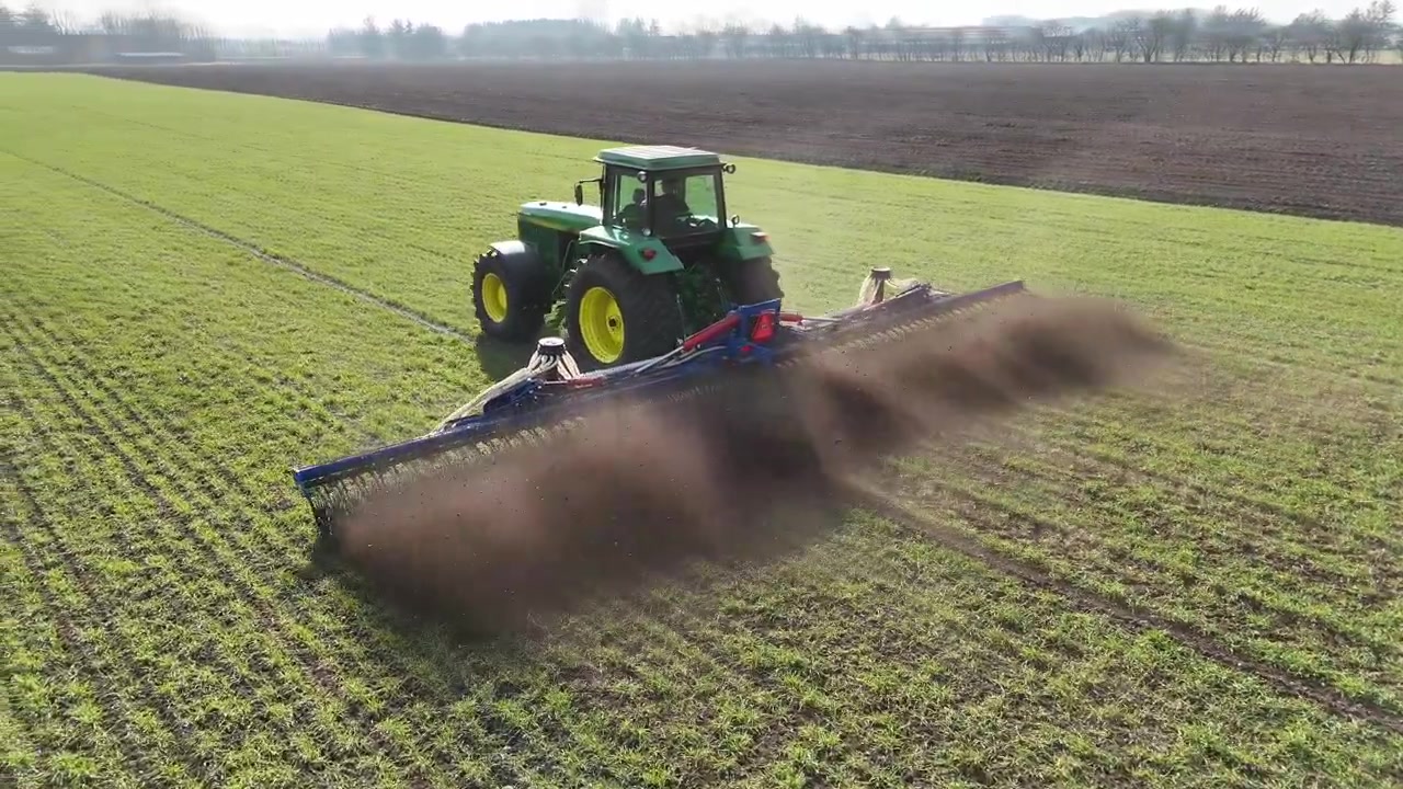 Riprese con drone seguono un trattore verde che coltiva un campo agricolo mentre la telecamera si muove intorno e rivela i macchinari in funzione da dietro