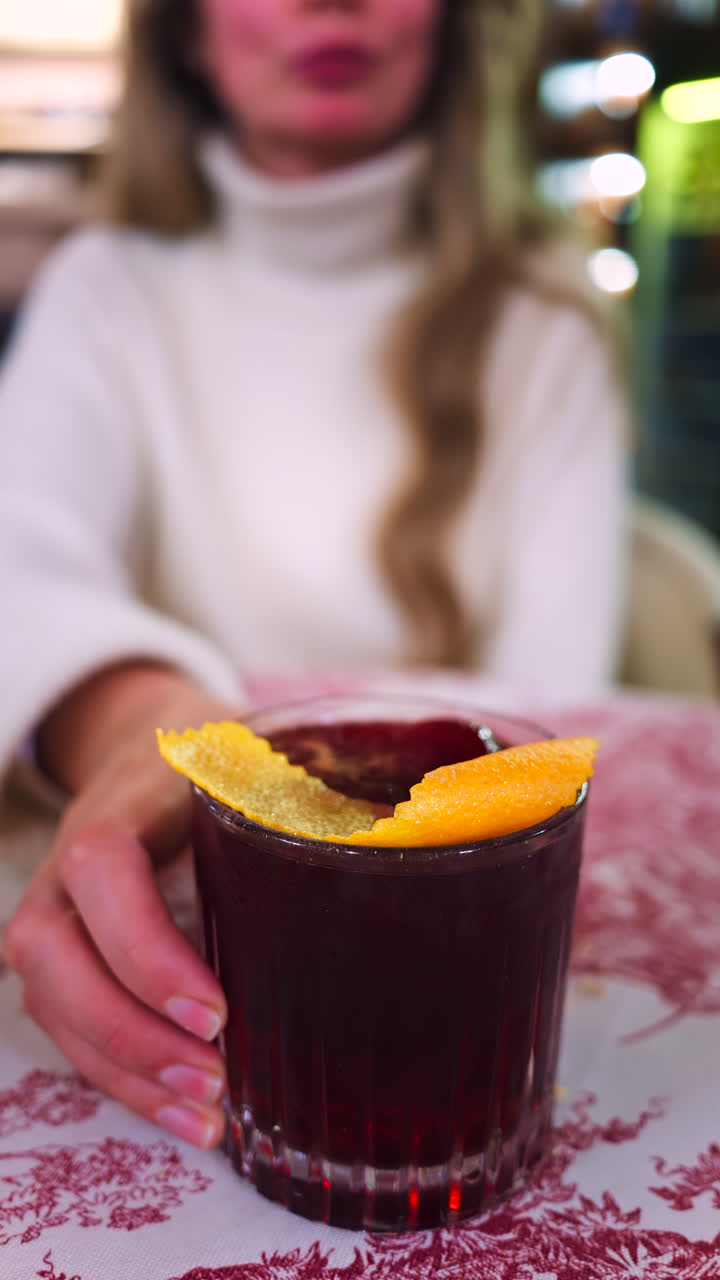 Close up of a woman holding a negroni cocktail on a red and white tablecloth at a restaurant. Vertical