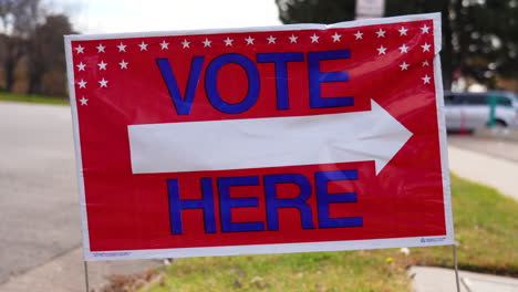 vote here sign arrow pointing left with people driving cars in background, close up