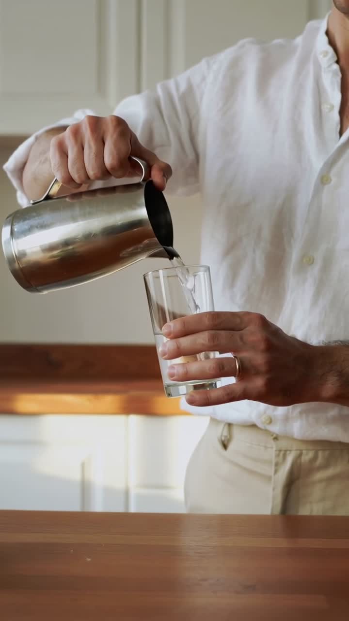 Vidéo verticale : Un homme en chemise blanche se penchant pour verser le contenu d'une carafe dans un verre sur le comptoir de la cuisine.