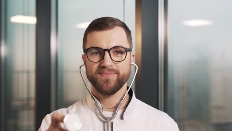 portrait of a male doctor in a white coat wearing a stethoscope and directing an earpiece towards the camera against the backdrop of panoramic windows in a modern new clinic