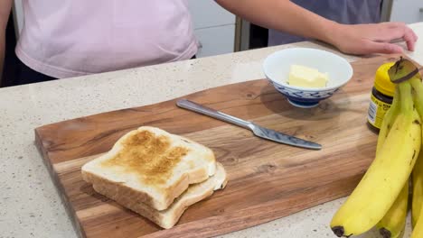 person removes toast from toaster, stacks slices on cutting board, and prepares to add butter and banana in a brightly lit kitchen setting