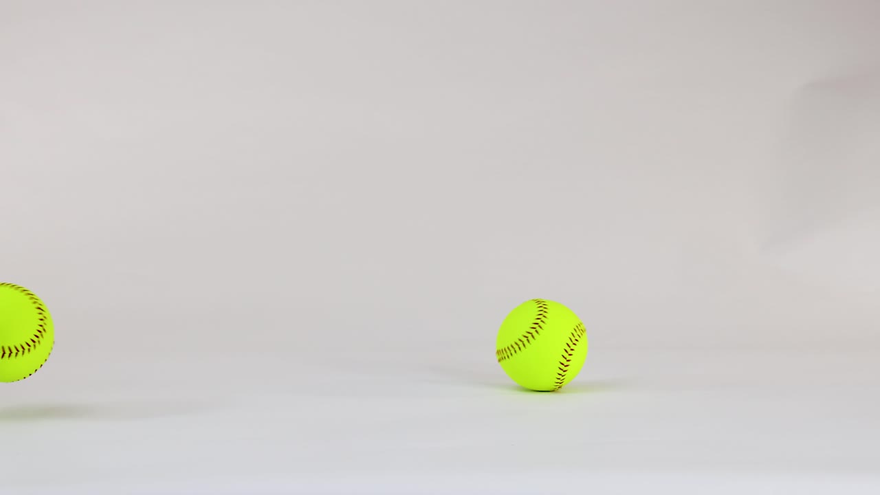 Una palla da softball giallo neon rimbalza e rotola sul pavimento di uno studio bianco con illuminazione soffusa