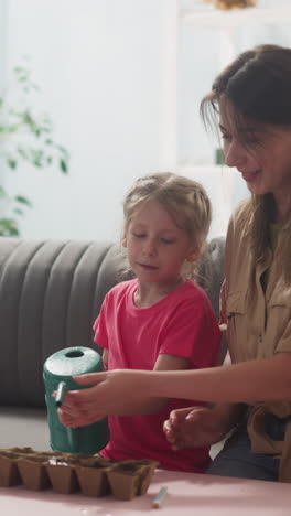 preschooler girl waters sowed seeds with watering can in seedling tray with mother and brother in living room slow motion. gardening hobby for whole family