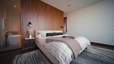 a luxury bedroom with white bedding and a wooden wall headboard and glass walls showing a free standing bathtub, wide push in shot at an angle