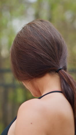 portrait back view of young woman stretching neck gently outdoors with blurred iron fence and trees in background, promoting relaxation and flexibility in natural setting