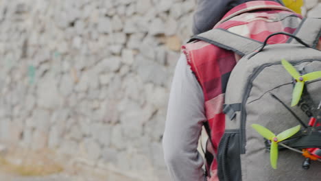 a man with a backpack behind him and a sports drone walks along a stone wall