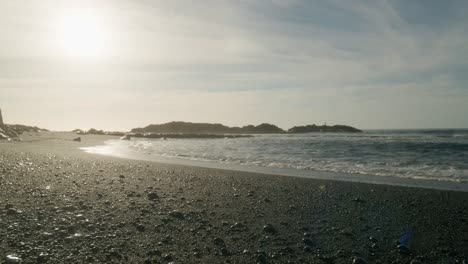 Tenerife-black-sand-beach-pebbles-sun-reflection-along-shore-clear-sky-low-angle,-Canary-Islands