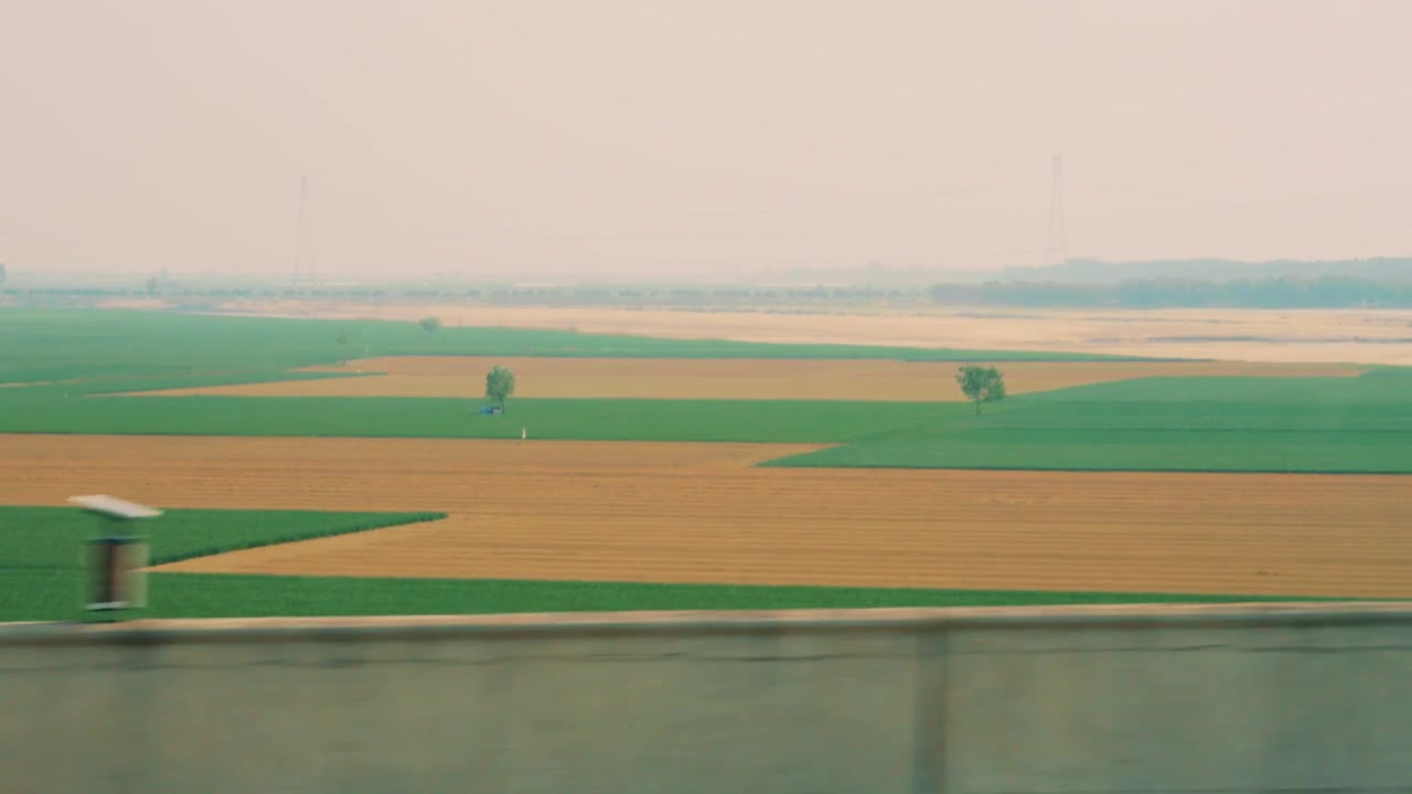 Una ripresa in movimento, con vista laterale, di campi agricoli altamente organizzati vicino a Xi'an. Il paesaggio presenta un distinto mosaico di terra verde e marrone, punteggiato da alberi solitari e linee di servizio.