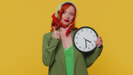 redhead young woman showing time on wall office clock, ok thumb up approve pointing finger at camera