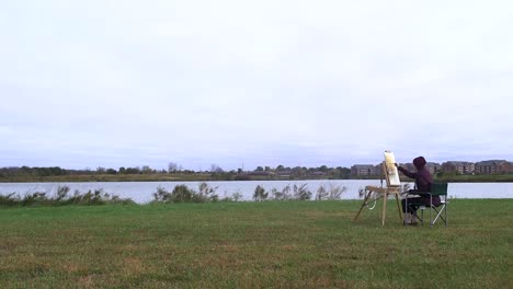 Young-Woman-Pints-Painting-Next-To-Park-Lake-on-Cloudy-Day-Wide-Shot