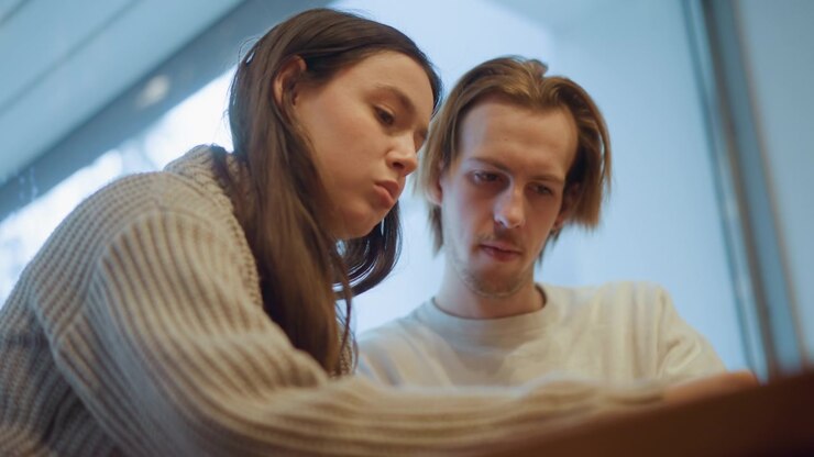Primo piano di due persone, una donna con un maglione a maglia beige e un uomo con una camicia chiara, concentrati su qualcosa insieme. Una luce naturale soffusa illumina la loro interazione all'interno, enfatizzando la calma.