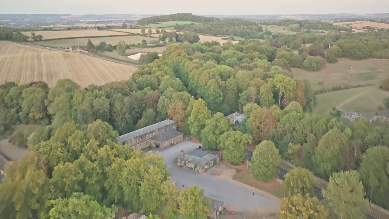 Ascending Aerial view of the round crenellated tower and adjacent stone house in Wentworth Castle Gardens, South Yorkshire, England, surrounded by autumn trees and historic parkland