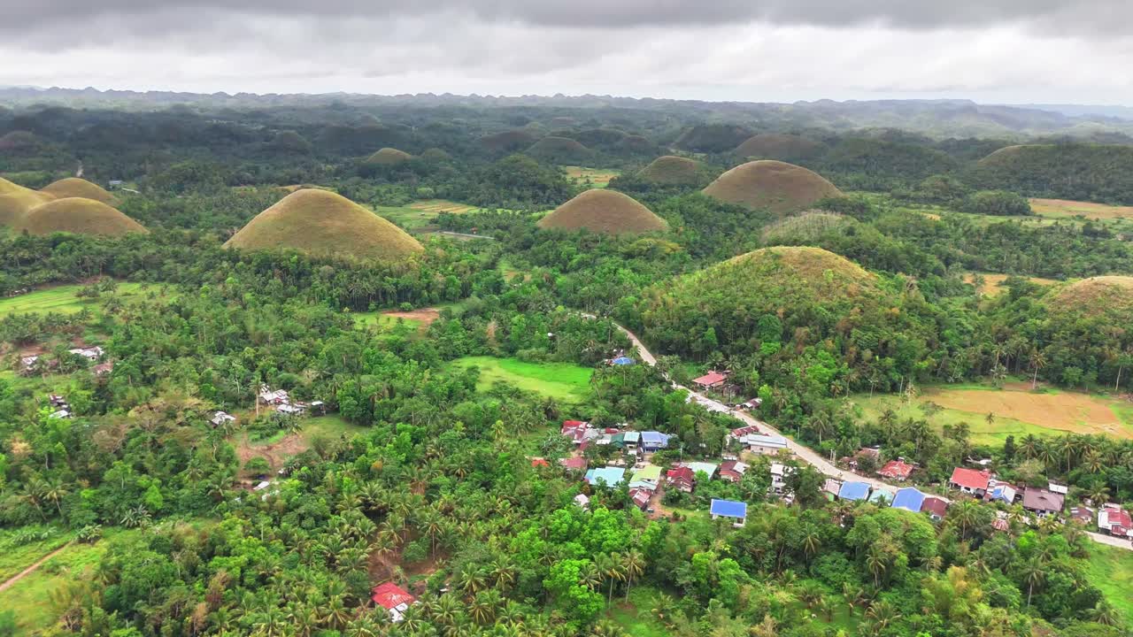 Lush green forest and rural village surround the iconic Chocolate Hills in Bohol, Philippines under a cloudy sky. Unique conical mounds dominate horizon, forming a natural wonder across the landscape