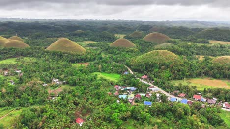 lush green forest and rural village surround the iconic chocolate hills in bohol, philippines under a cloudy sky. unique conical mounds dominate horizon, forming a natural wonder across the landscape