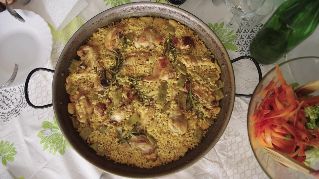 Vista dall'alto di una tradizionale paella valenciana spagnola su una tavola familiare apparecchiata, mentre delle mani usano utensili di legno per mescolare un'insalata fresca