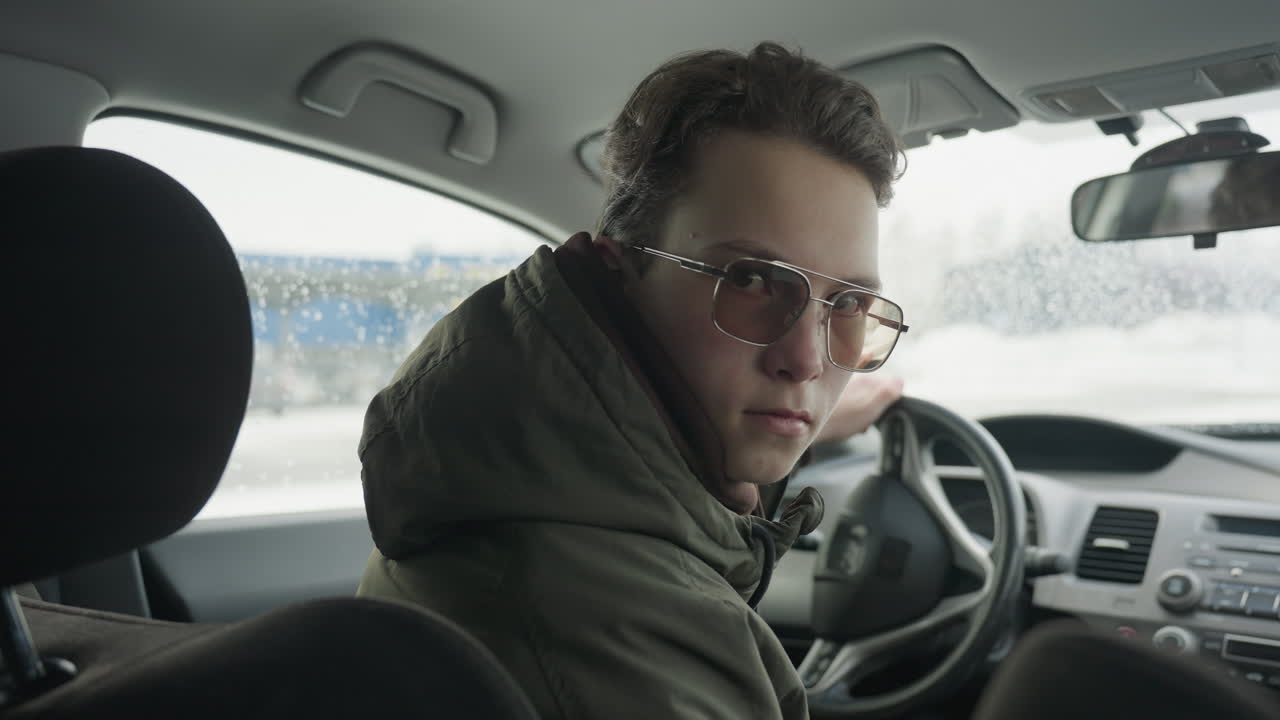 young teenager wearing winter jacket and sunglasses turns back from driver seat to look directly at camera inside parked car with cold snowy exterior and blurred city buildings visible through window