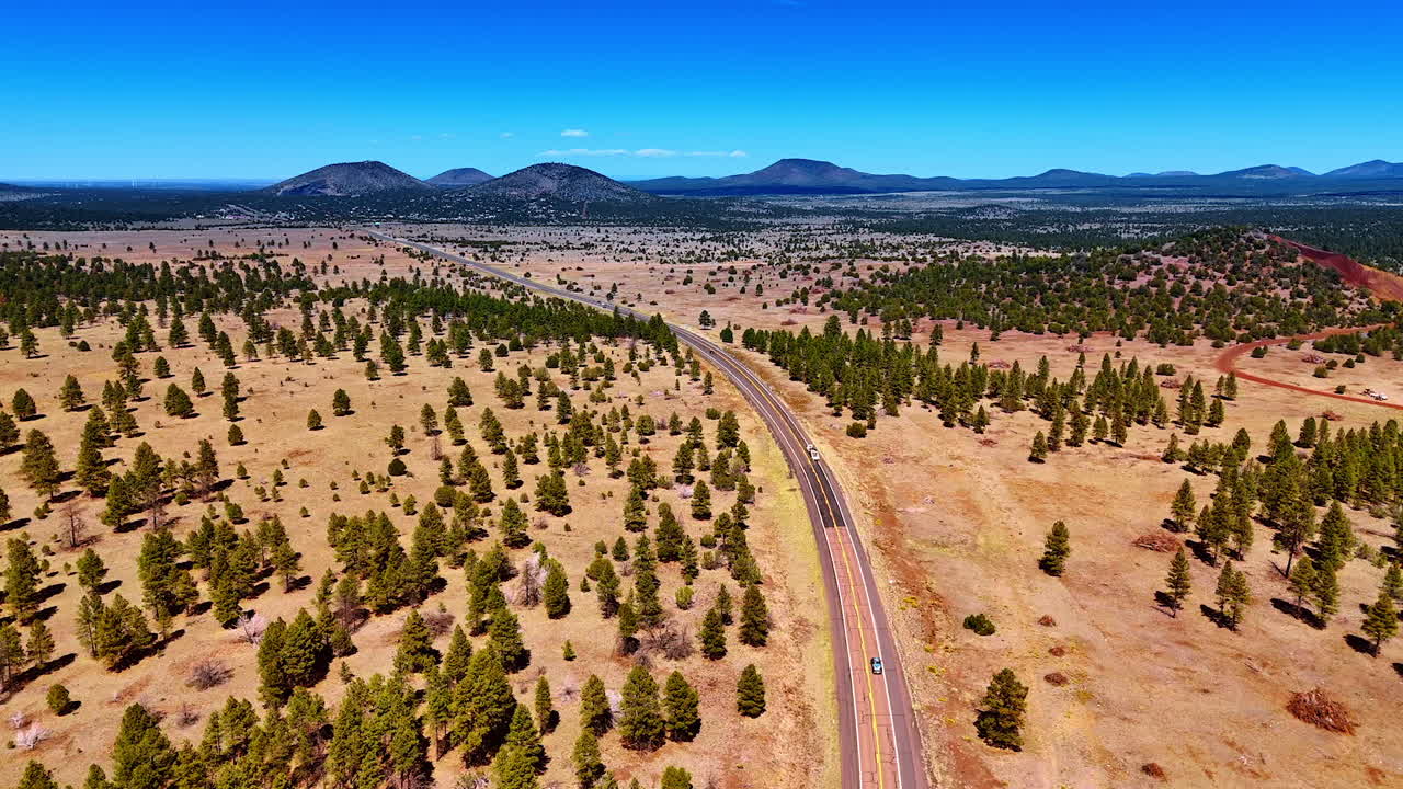Following vehicles riding by the highway along the dry landscape. Sparse pine tree woods grow around. Mountains at backdrop. Arizona, USA