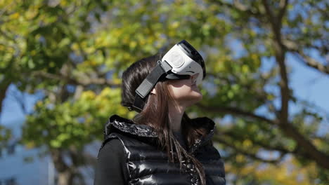 young dark-haired woman in warm black waistcoat wearing virtual reality glasses in early autumn park