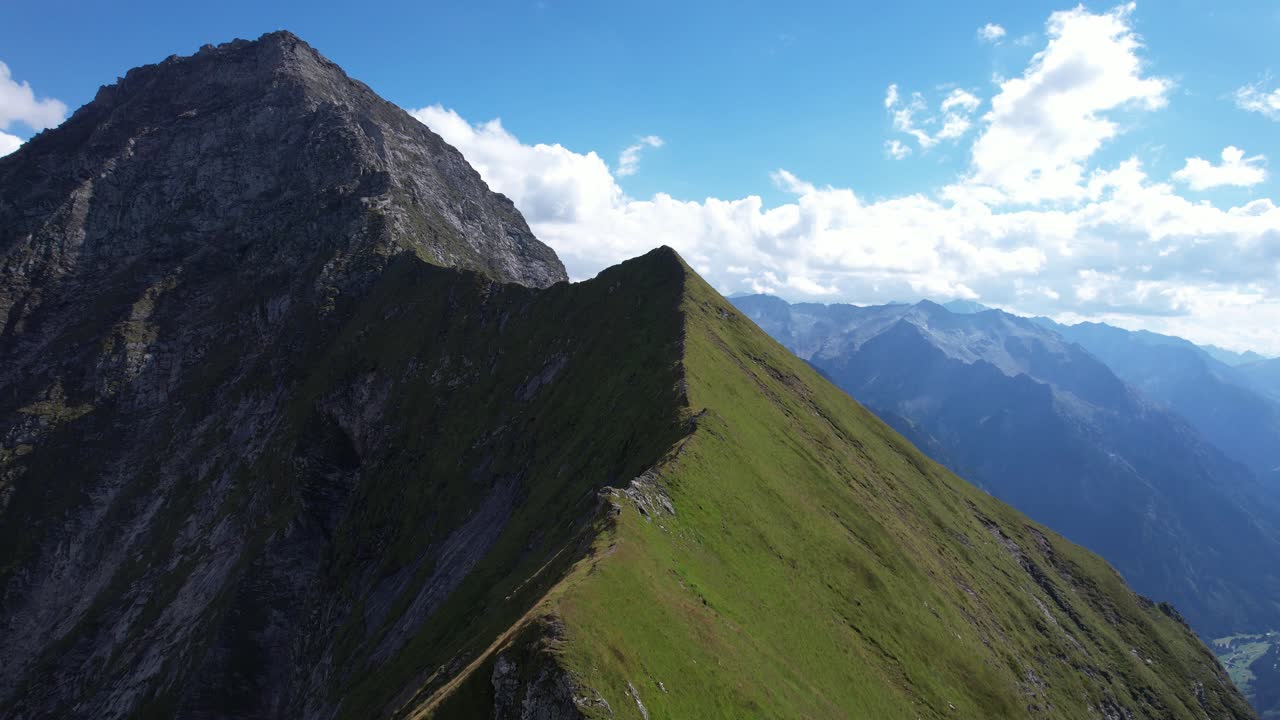 una cresta aguda y cubierta de hierba asciende a un pico rocoso bajo un cielo azul brillante, con majestuosas cadenas montañosas en la distancia, capturando la belleza escarpada de los alpes austriacos en un pequeño pueblo llamado mayrhofen.