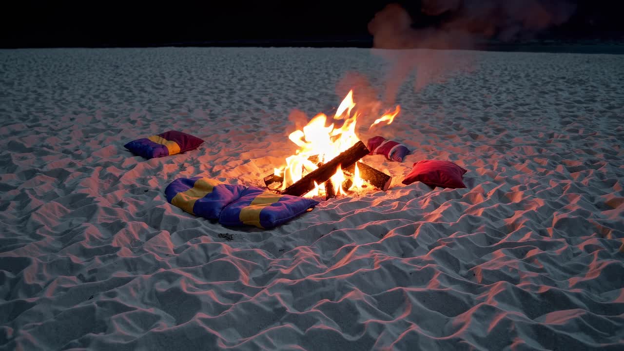 Bonfire burning brightly on a sandy beach at night, casting a warm glow over colorful pillows arranged around it, creating a cozy and inviting atmosphere for relaxation and enjoyment
