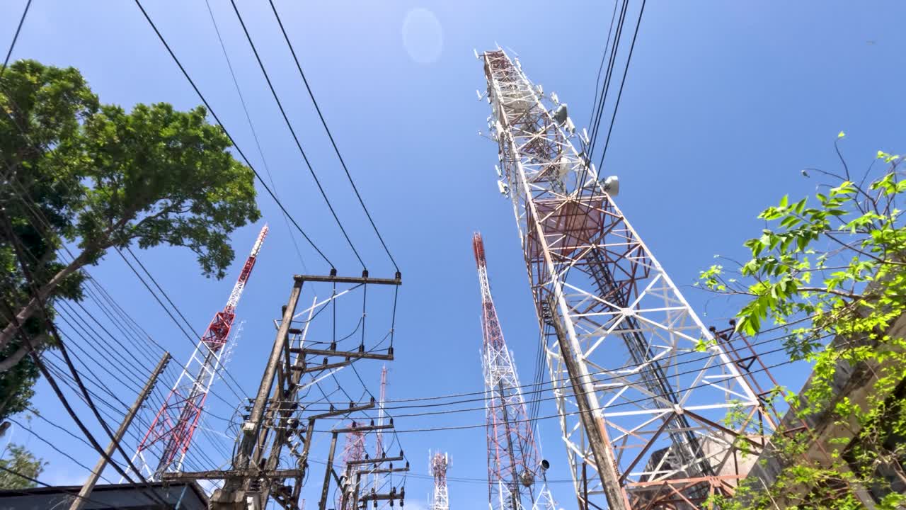 Low-angle camera pans upward past cell towers, power cables, and trees under bright daylight sky