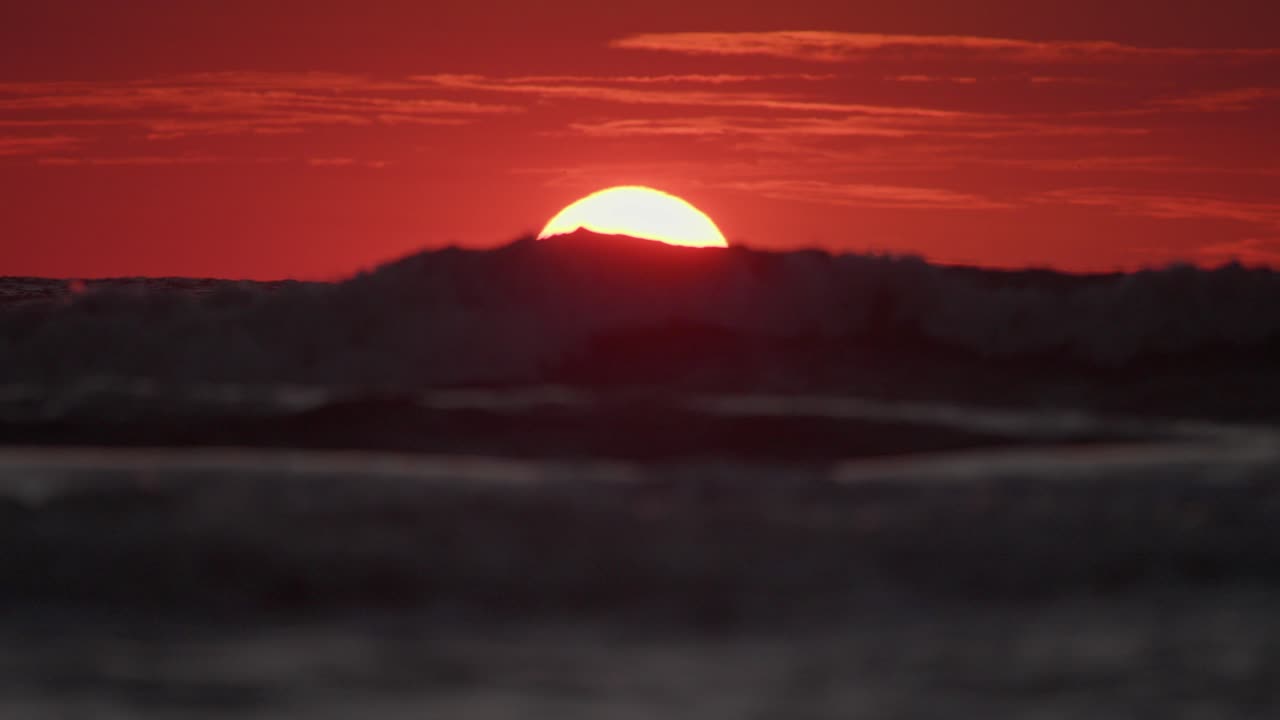 Meereswellen rollen über das Wasser, während die gelbe Sonne bei rotem Himmel unter dem Horizont versinkt, Teleaufnahme statisch.