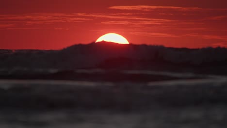 ocean waves roll across water as yellow ball of sun sets below horizon with red sky, telephoto static
