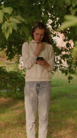 lady in casual clothing stands under tree, deeply focused on phone with hand on hood collar, sunlight filtering through leaves, with gentle greenery in background swaying slightly