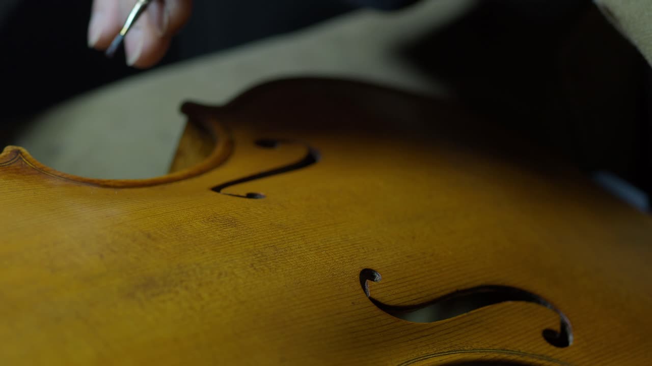 Hands of a luthier precisely darkening violin edges, simulating natural wear and aging on the handcrafted instrument on edges and purfling, with finger and paintbrush traditional tecqnique