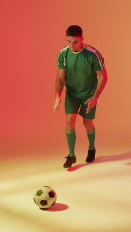 vertical video of caucasian male soccer player kicking football over neon pink lighting