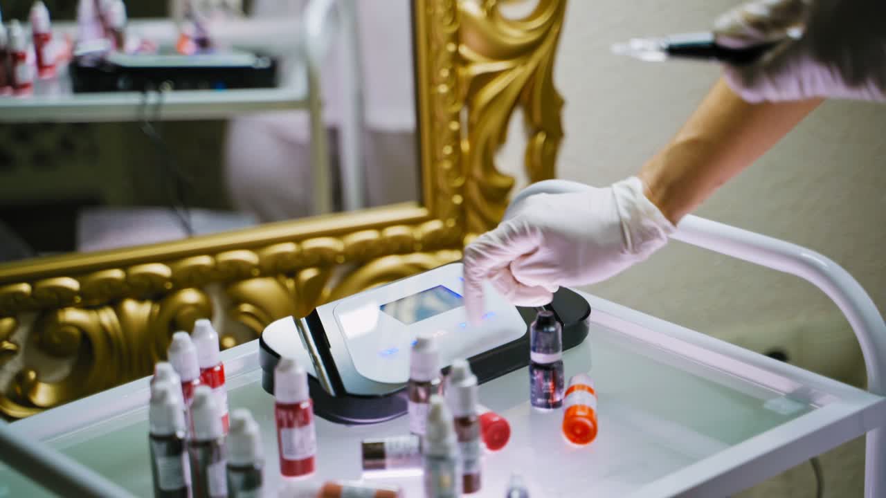 Master in white gloves turns on an electric digital permanent makeup machine on the table surrounded by jars with pigments in front of a mirror in a beauty salon. Preparation for the procedure.