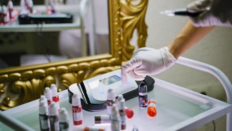 master in white gloves turns on an electric digital permanent makeup machine on the table surrounded by jars with pigments in front of a mirror in a beauty salon. preparation for the procedure.