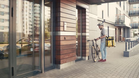 il corriere per la consegna di cibo che indossa uno zaino termico parcheggia la sua bicicletta accanto all'ingresso di un edificio per effettuare una consegna per clienti e clienti