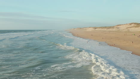 Olas-Rompiendo-En-La-Costa-De-Una-Hermosa-Playa-A-La-Luz-De-La-Tarde-Con-Gente-Divirtiéndose,-ángulo-Alto-Aéreo-Dando-Vueltas-Lentamente