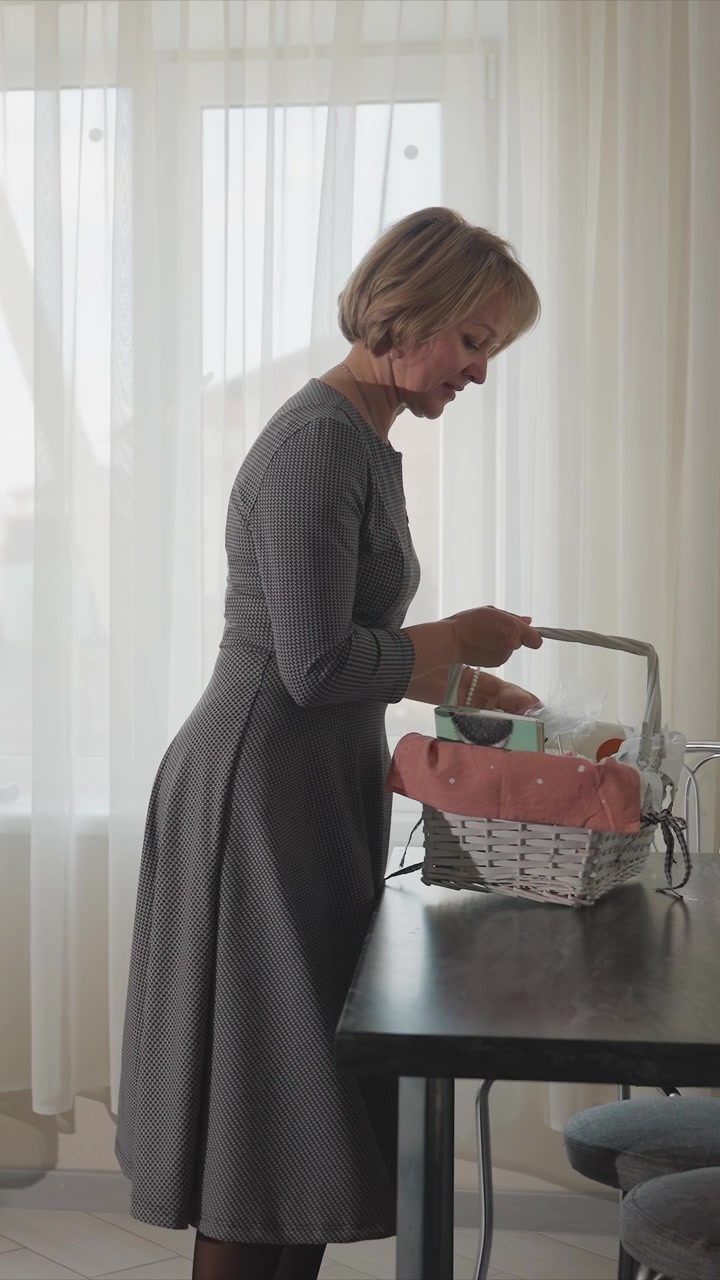 Close-up de uma mulher colocando itens de uma cesta de vime sobre uma mesa de madeira escura, em frente a espelhos hexagonais em uma parede cinza texturizada. Uma pequena planta em vaso está por perto, e a luz natural suave preenche o ambiente.