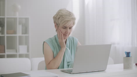 Una-Mujer-Anciana-Está-Trabajando-Con-Una-Computadora-Portátil-En-Casa-Sintiendo-Un-Dolor-De-Muelas-Agudo-Tocando-La-Mejilla-Y-Haciendo-Una-Mueca-De-Dolor