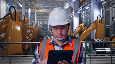 close up of asian male engineer with safety helmet working on a tablet while standing with industrial robotic arms working