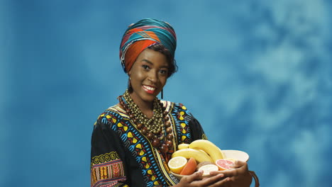 joven afroamericana alegre con turbante y ropa tradicional sonriendo alegremente a la cámara mientras sostiene un plato con frutas