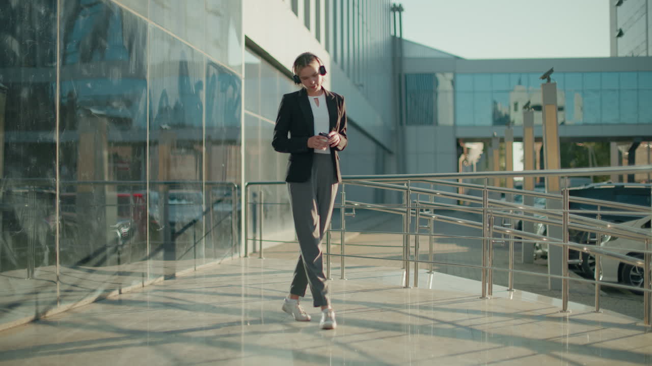Woman in professional outfit enjoying music while holding beverage, dancing casually on polished walkway with parked cars and glass building reflecting urban surroundings