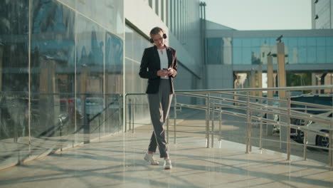 woman in professional outfit enjoying music while holding beverage, dancing casually on polished walkway with parked cars and glass building reflecting urban surroundings
