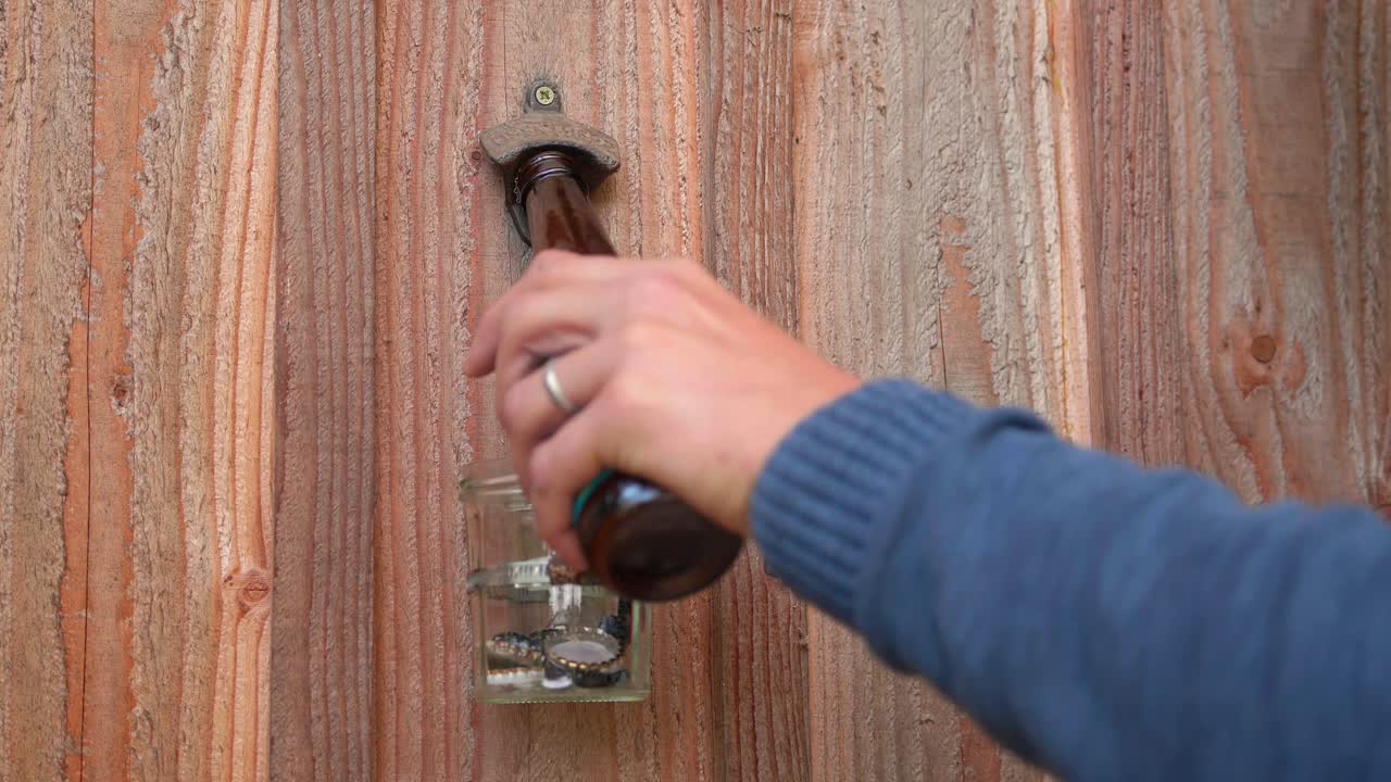 Una mano usa un apribottiglie in metallo montato su un pannello di legno per rimuovere il tappo da una bottiglia di vetro marrone. Primo piano.