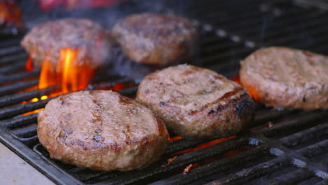 multiple meat burgers cooking on bbq fire flame grill with chef flipping hamburgers in background. barbecue summer seasonal cooking food.