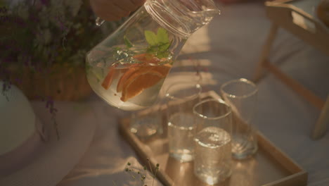 close-up of hand pouring refreshing infused water from pitcher with orange slices and mint into glasses on wooden tray, under warm sunlight, with partial view of picnic table