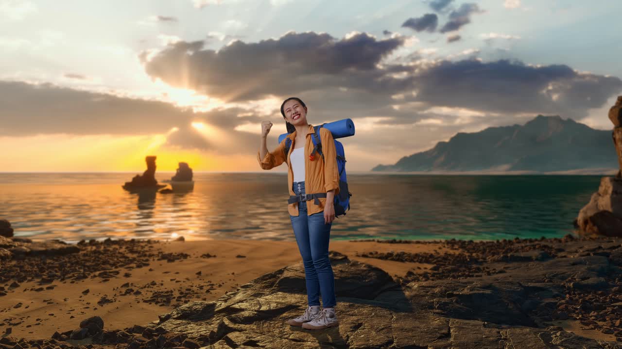 Inquadratura laterale a figura intera di escursionista asiatica con zaino da alpinismo che urla celebrando il successo mentre si trova in piedi sulla spiaggia durante il tramonto