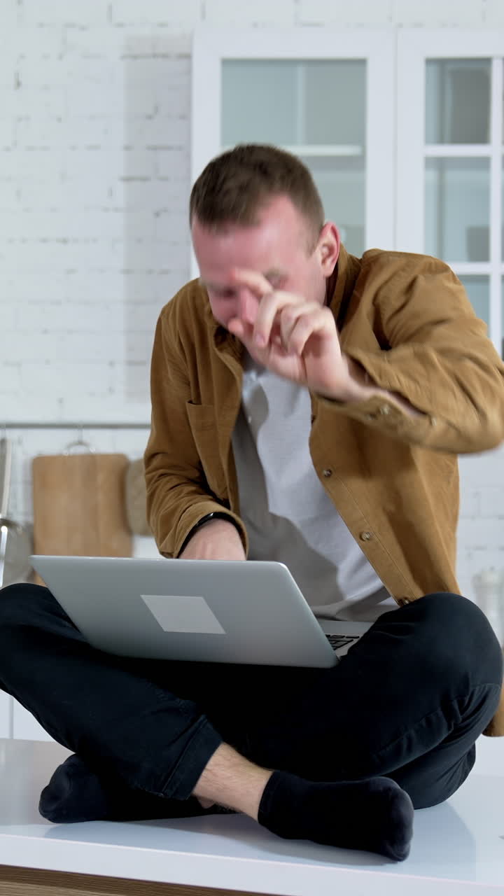 Emotional young man playing games at home. Funny man pointing with finger on camera while sitting on the kitchen table with a laptop. Vertical video