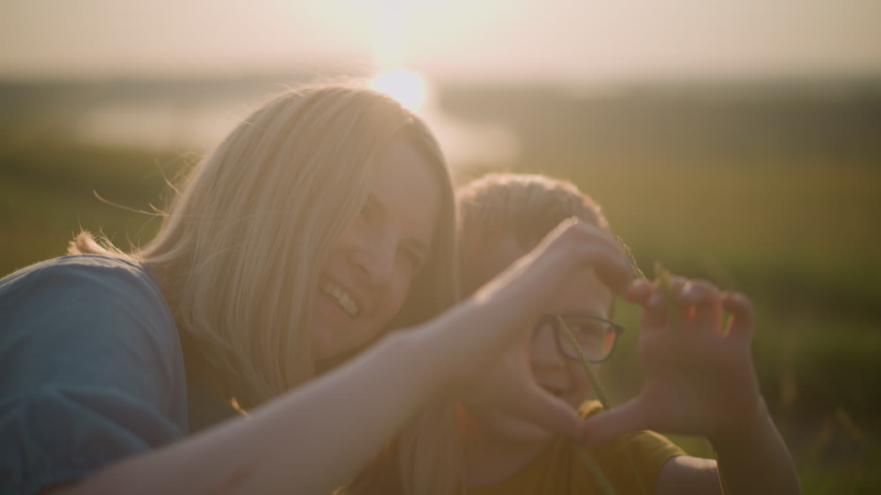 아름답게 흐릿한 이미지는 어린 소년과 여자의 감동적인 순간을 포착합니다. 머리가 함께 붙어, 웃으며 조용한 일몰 동안 손으로 심장 모양을 만니다.