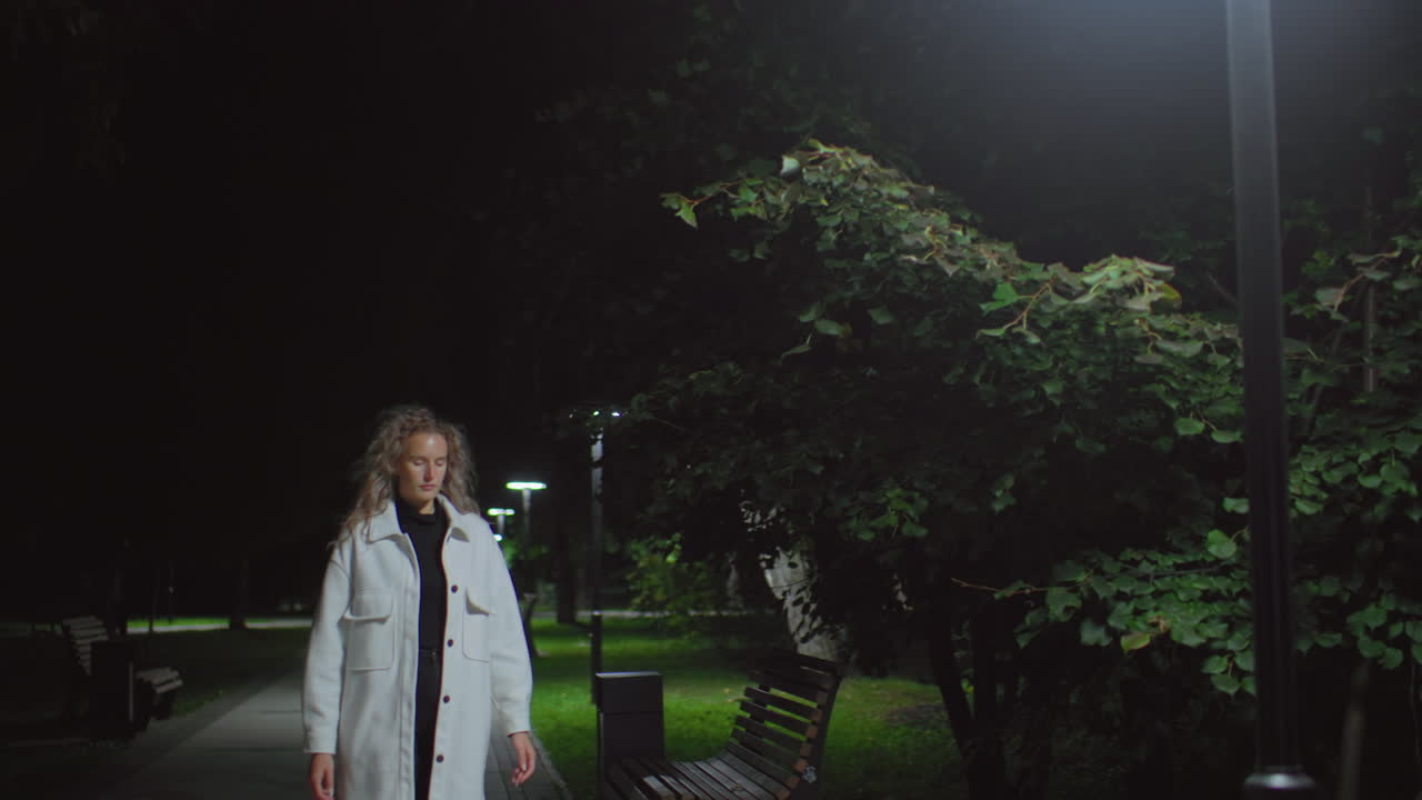 Young girl in white coat walks under dim street light on quiet path at night, benches lined up, faint figures visible behind, capturing calm ambiance and subtle tension in dark environment
