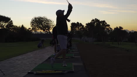 Grupo-De-Golfistas-En-El-Campo-De-Prácticas-Golpeando-Pelotas-Con-El-Amanecer-Detrás-De-Ellos